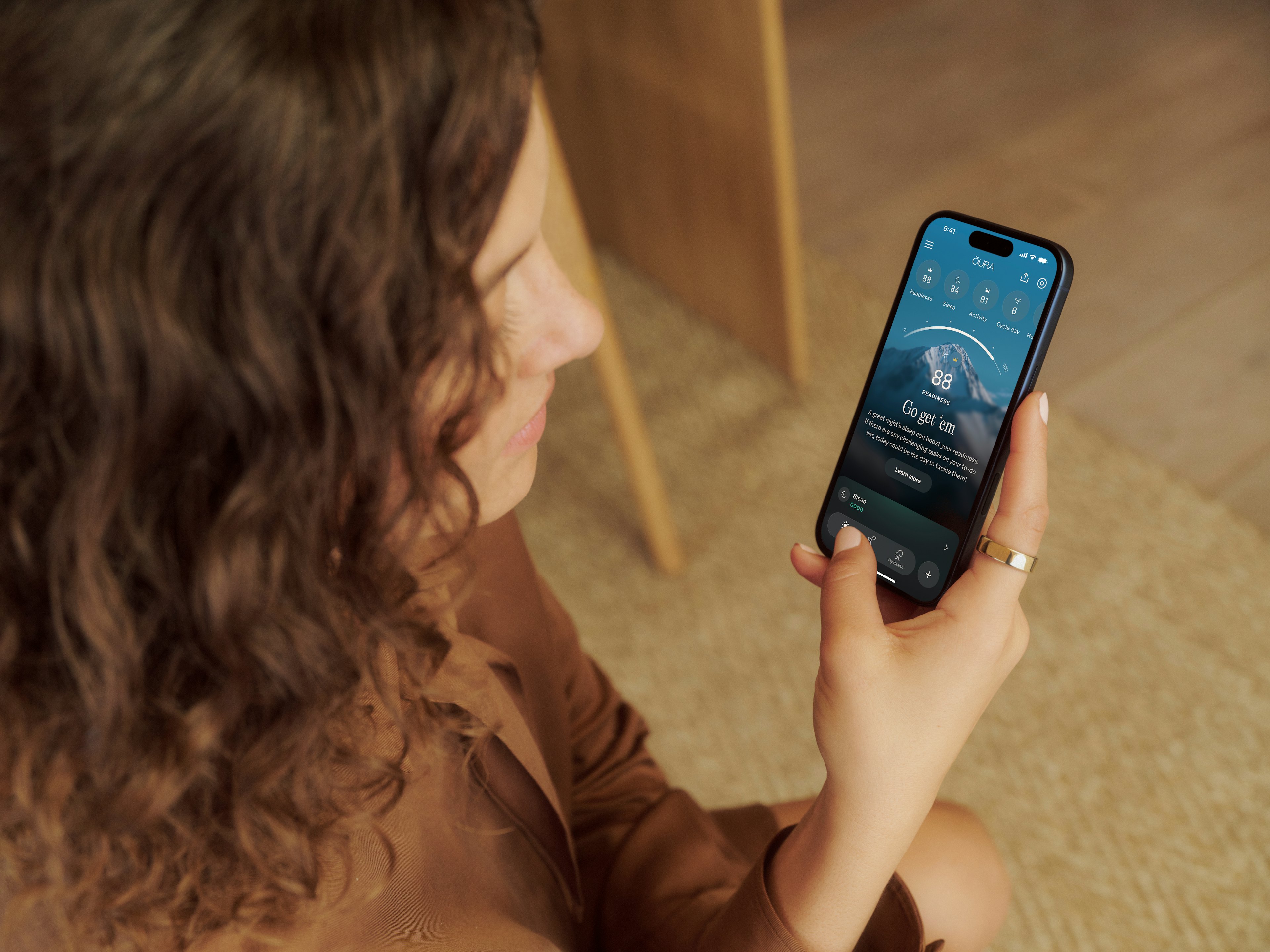 一位卷发女子手持智能手机，佩戴 Oura Ring 5，正在查看 Oura App。