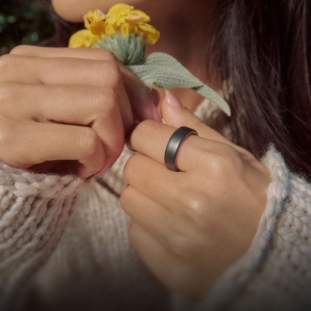 一個人手上拿著一朵花，手指上戴著 Stealth 色 Oura Ring 5 的特寫