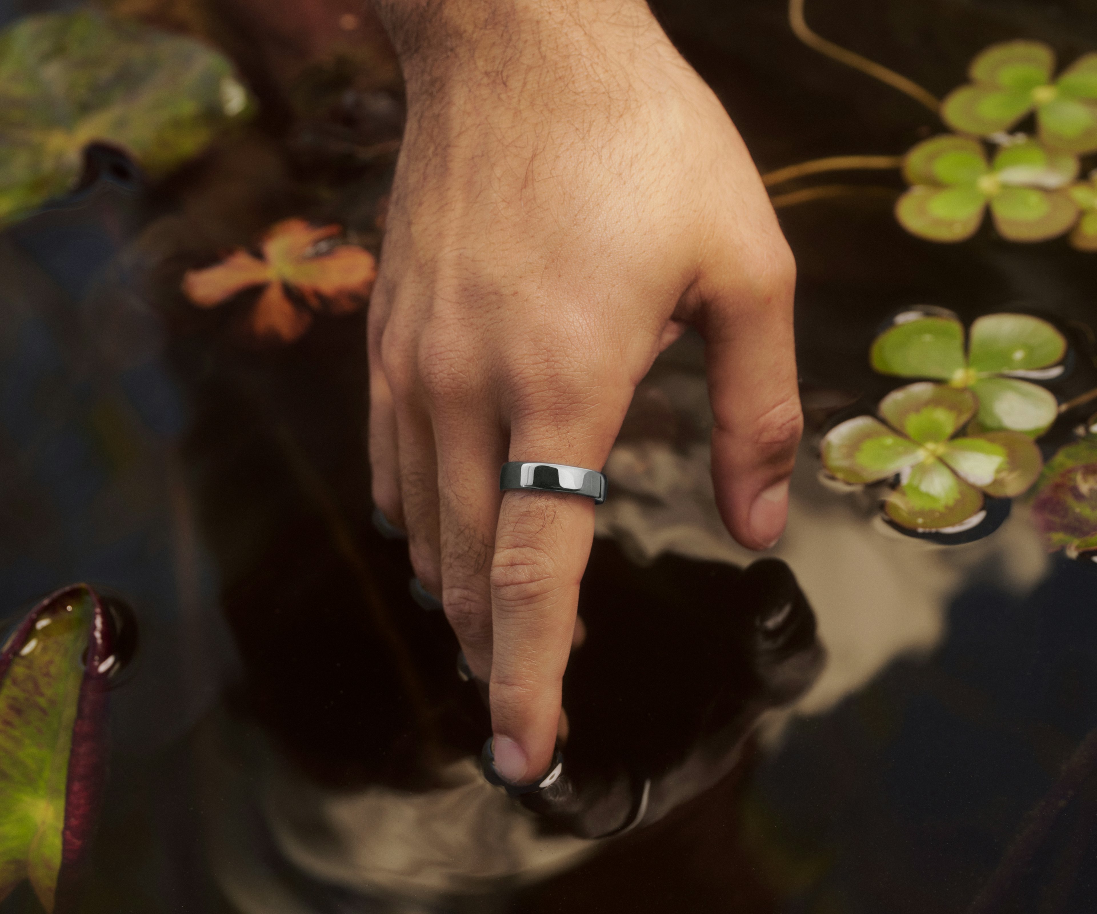 特写：一个人单手轻触景观池塘水面，手指上佩戴着 Silver 饰面的 Oura Ring 5