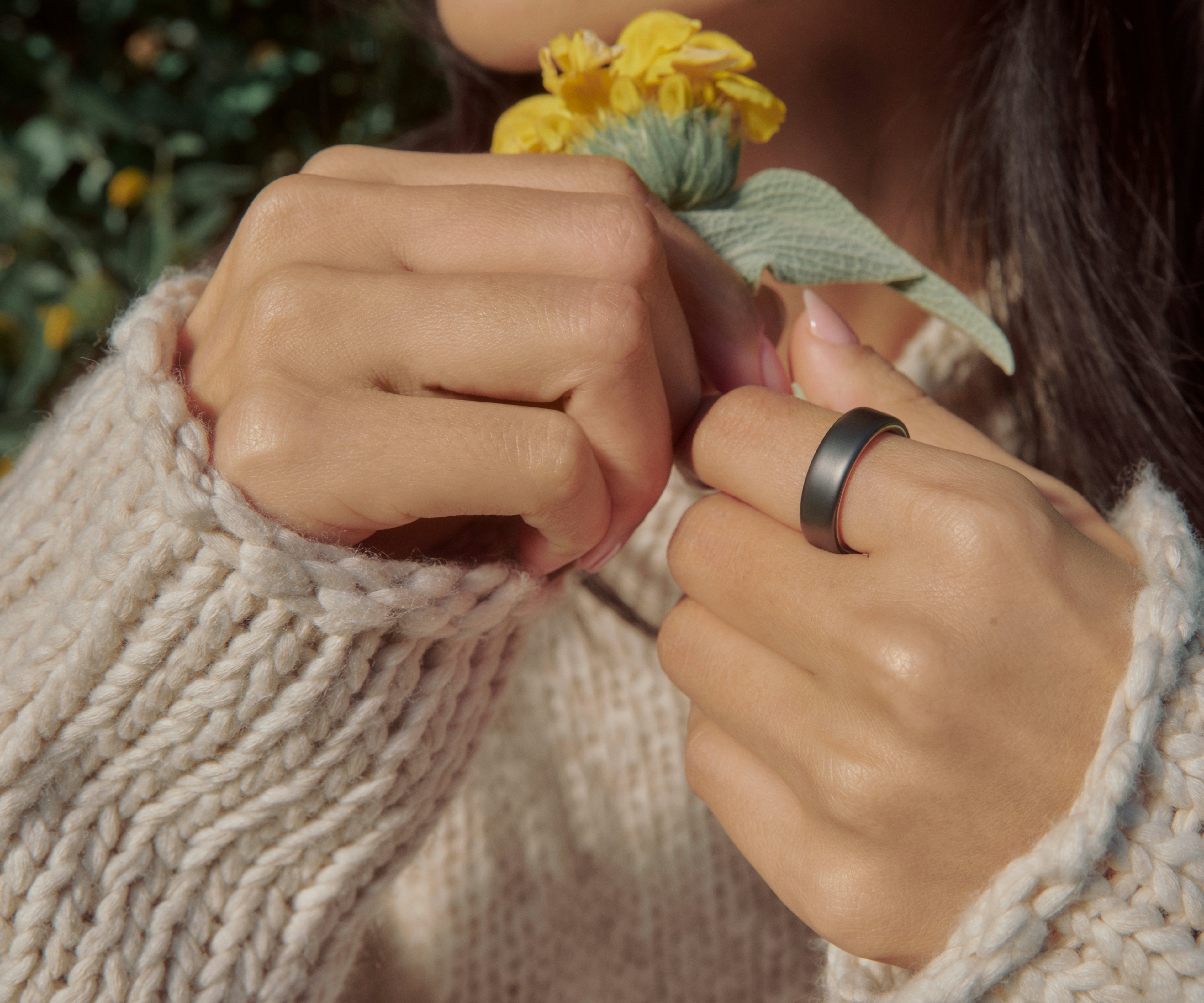 特写：一个人双手轻拈花朵，手指上佩戴着 Stealth 饰面的 Oura Ring 5